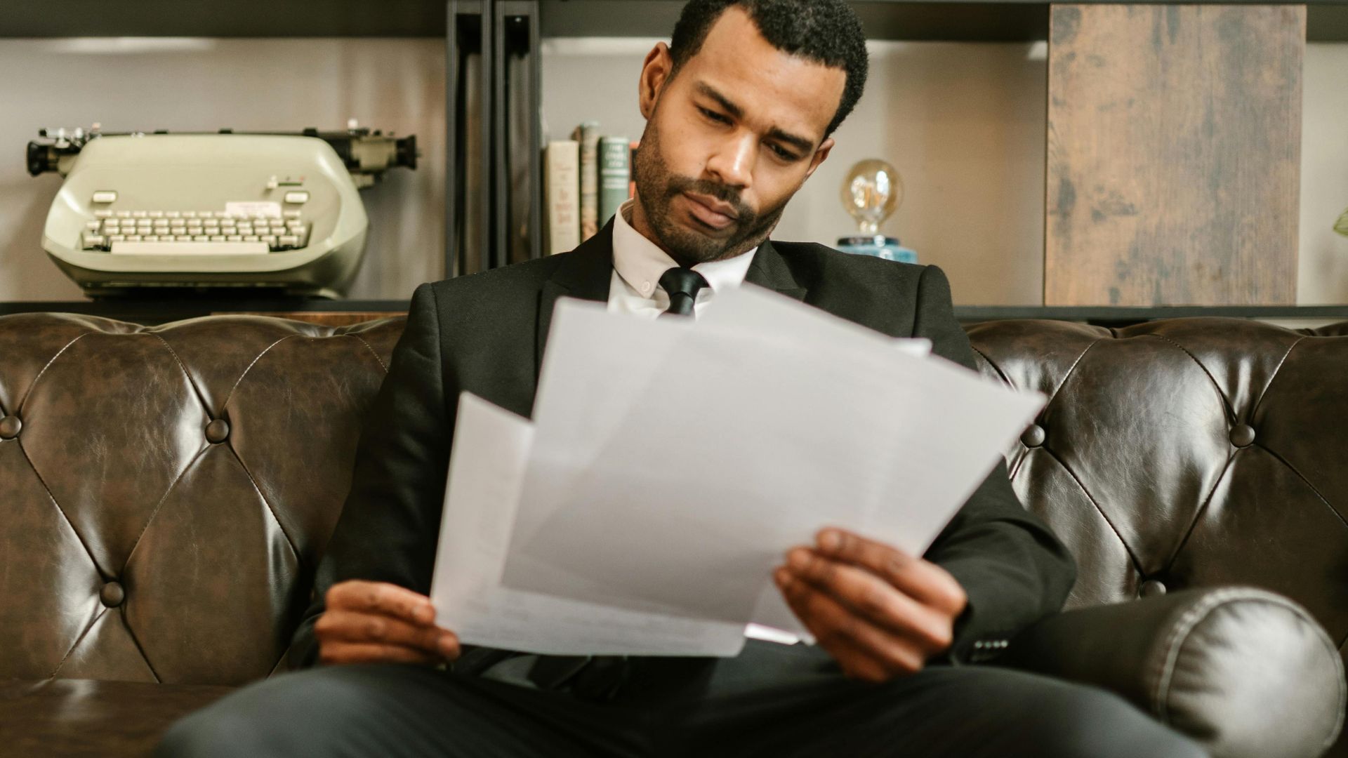 Adult man in a suit reading paperwork on a leather couch in a stylish office setting.