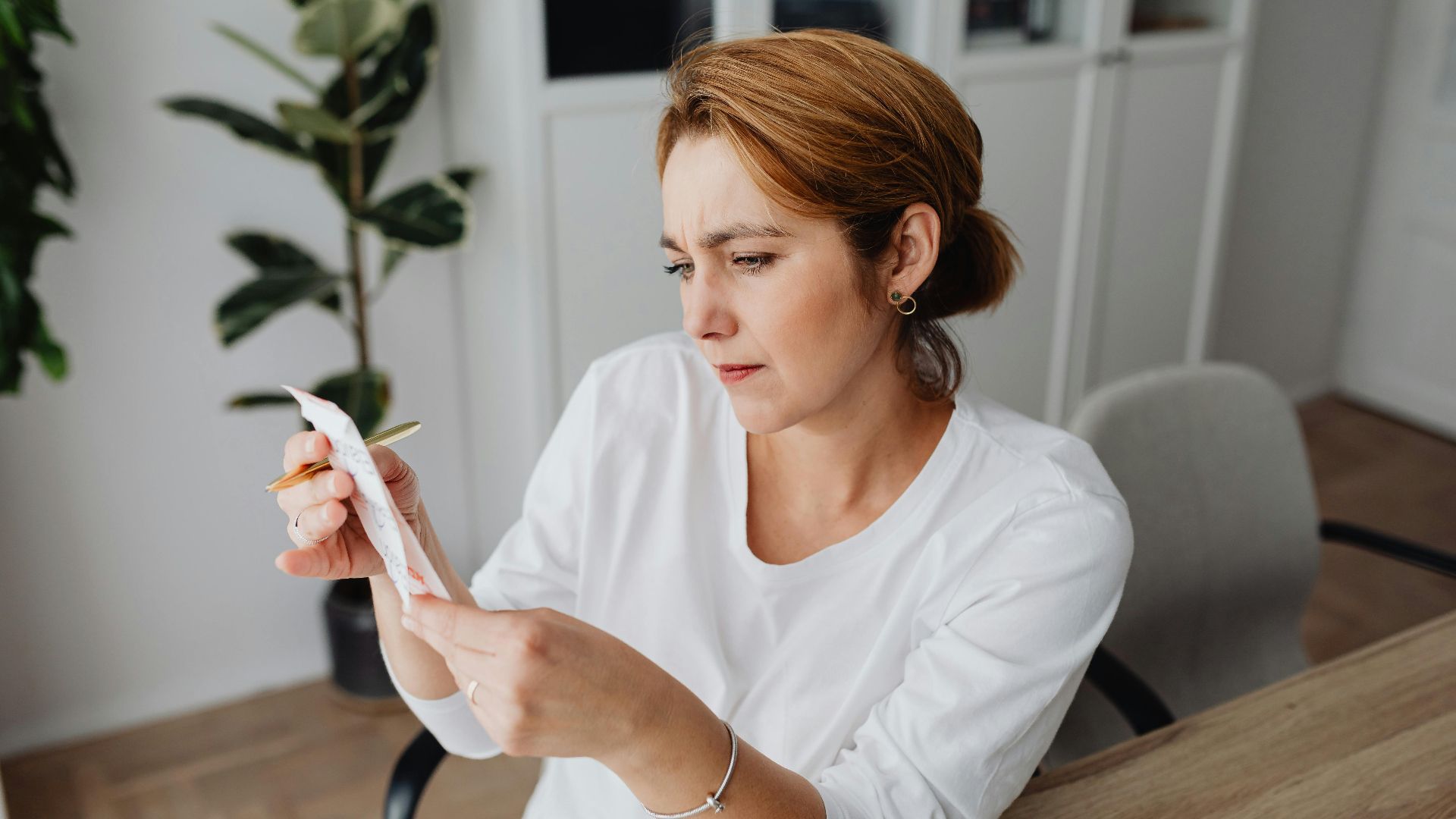 A concerned adult woman examines a receipt while sitting at her office desk, indicating financial scrutiny.