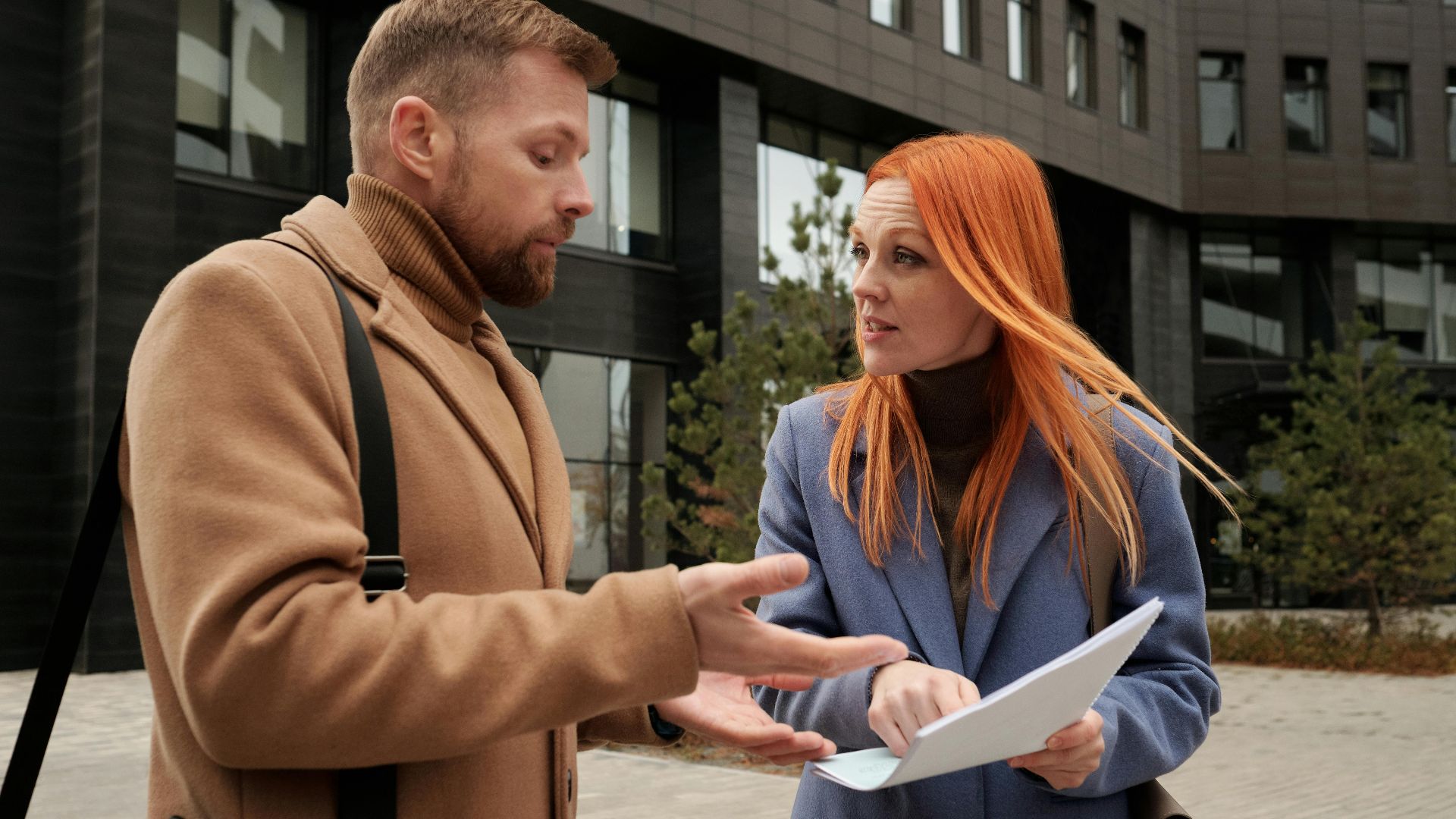 A man and woman in coats discussing documents outside a modern building.
