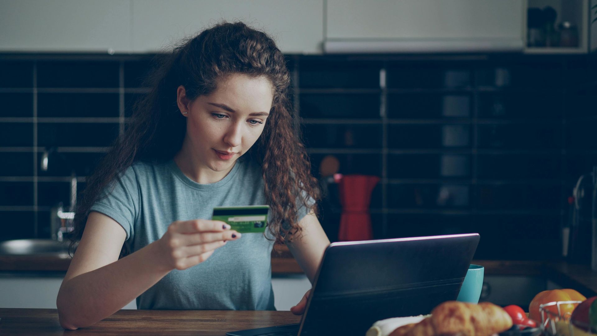 Young woman using a credit card to shop online with a laptop in a kitchen setting.