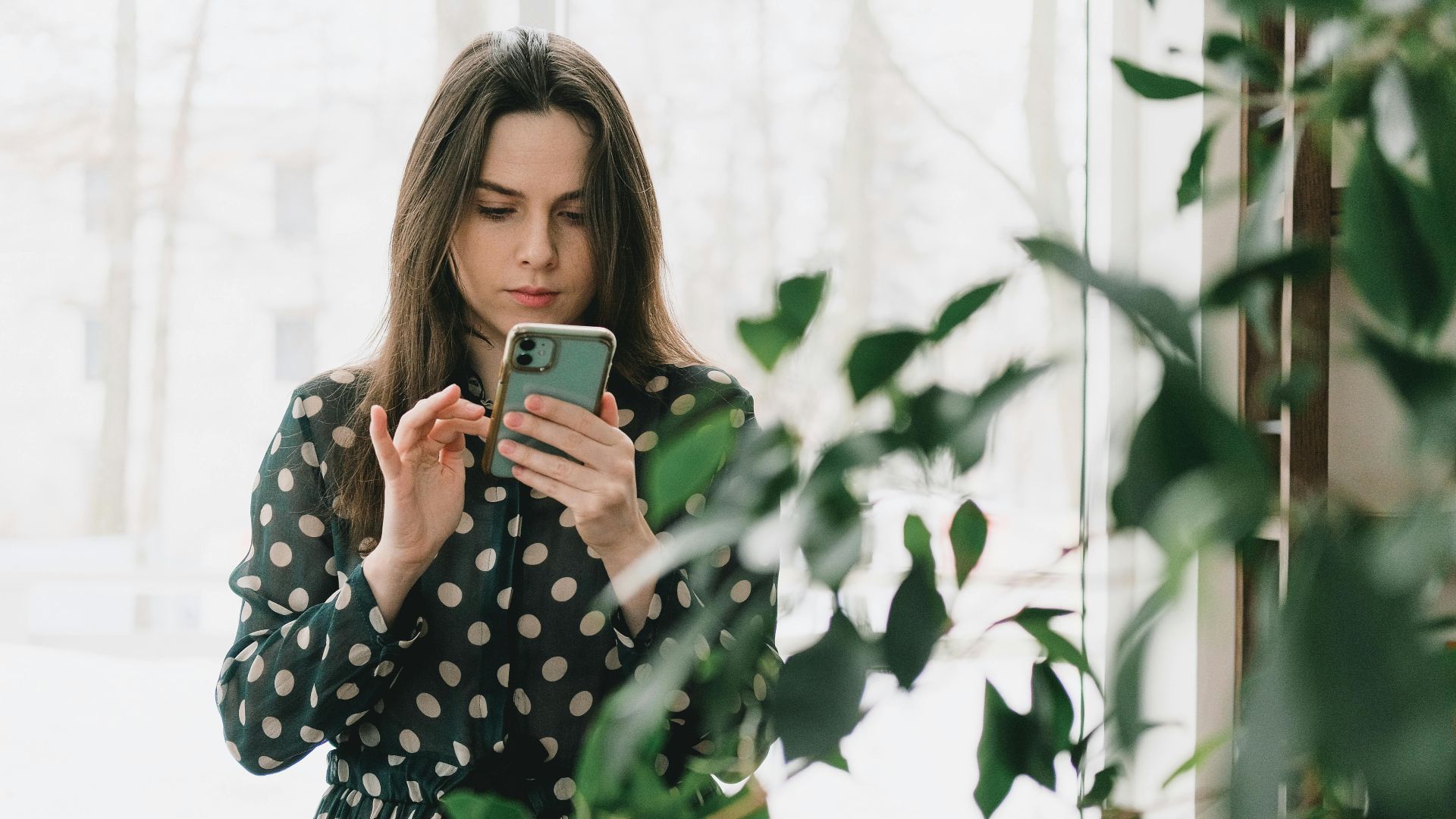 Woman in polka dot dress using smartphone indoors, surrounded by plants.