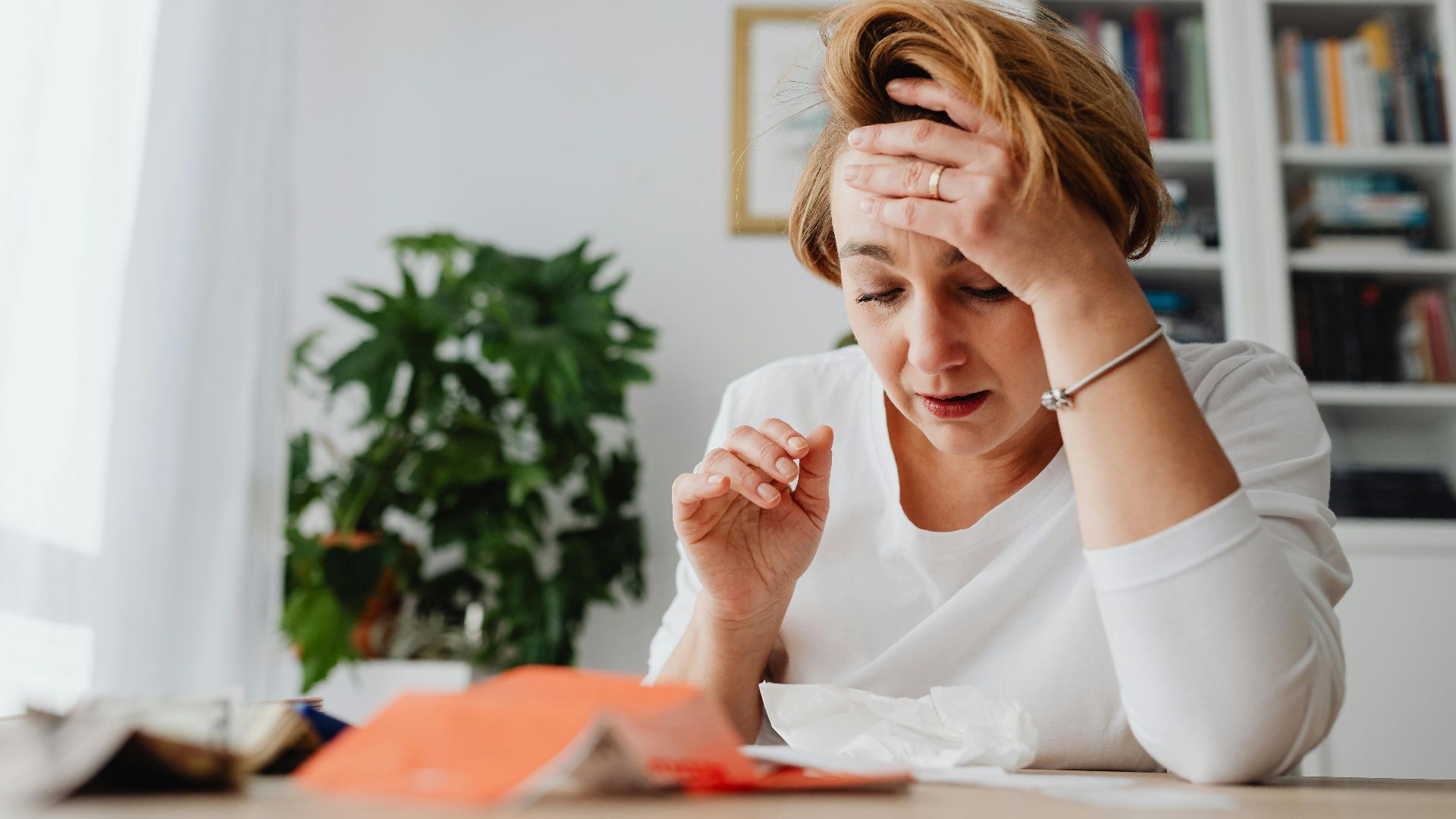 A stressed woman in a white shirt holds her head at a desk cluttered with papers, indoors.