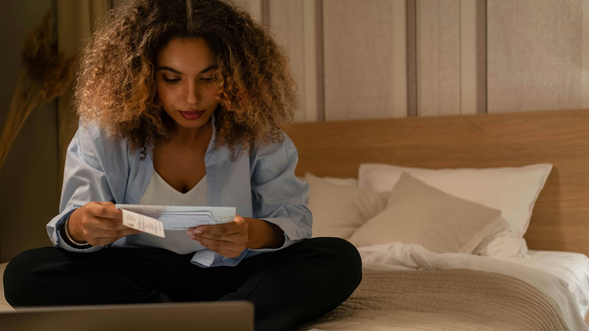 Woman sitting on bed reading documents with laptop, highlighting home office setting.