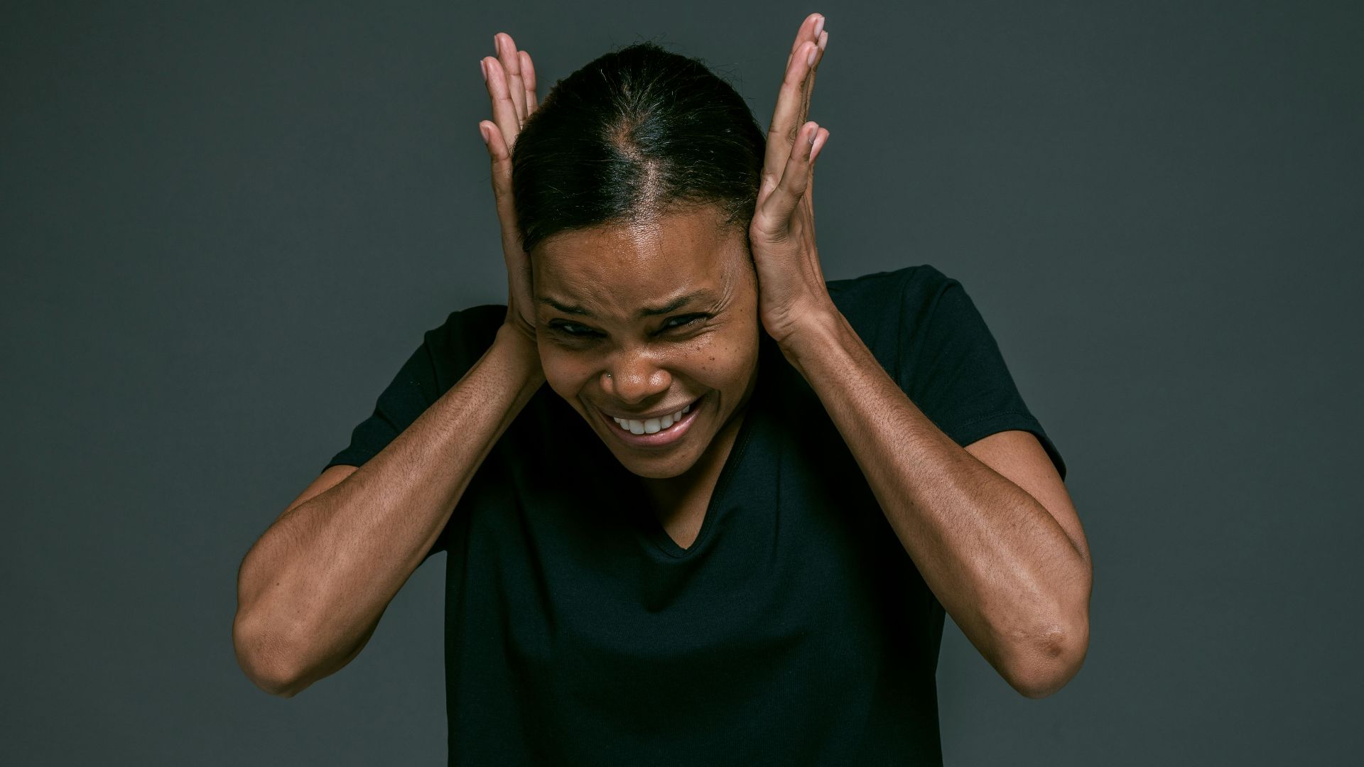Portrait of an adult woman covering ears, looking irritated, against a dark background.