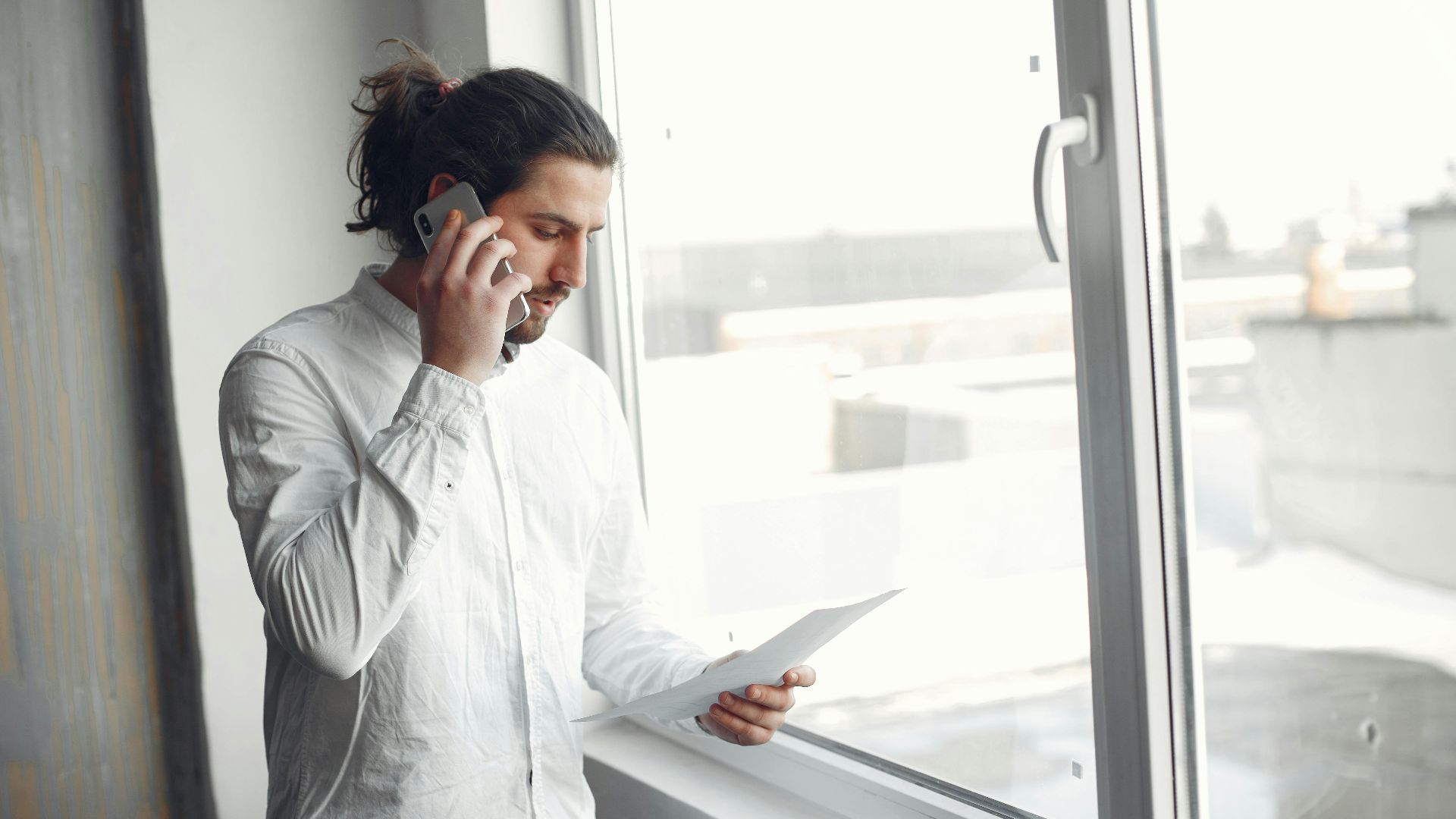 Young man in white shirt, on phone call holding a document, standing by a large window.