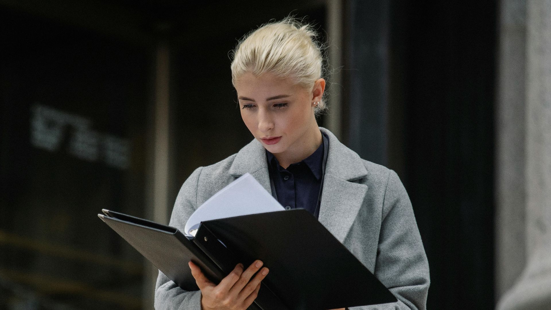 Focused young businesswoman in formal clothes standing outside modern building and reading notes in folder
