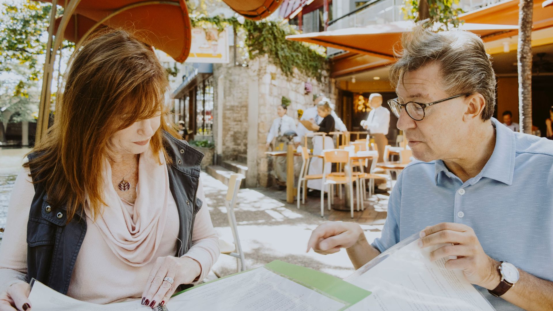 man and woman sitting down near table while reading outside cafe during daytime