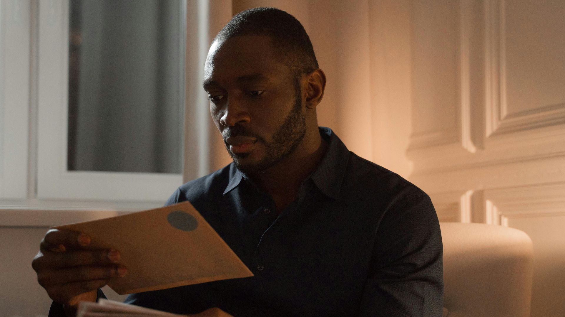 A man with a mustache reads letters while sitting in a warmly lit home interior.