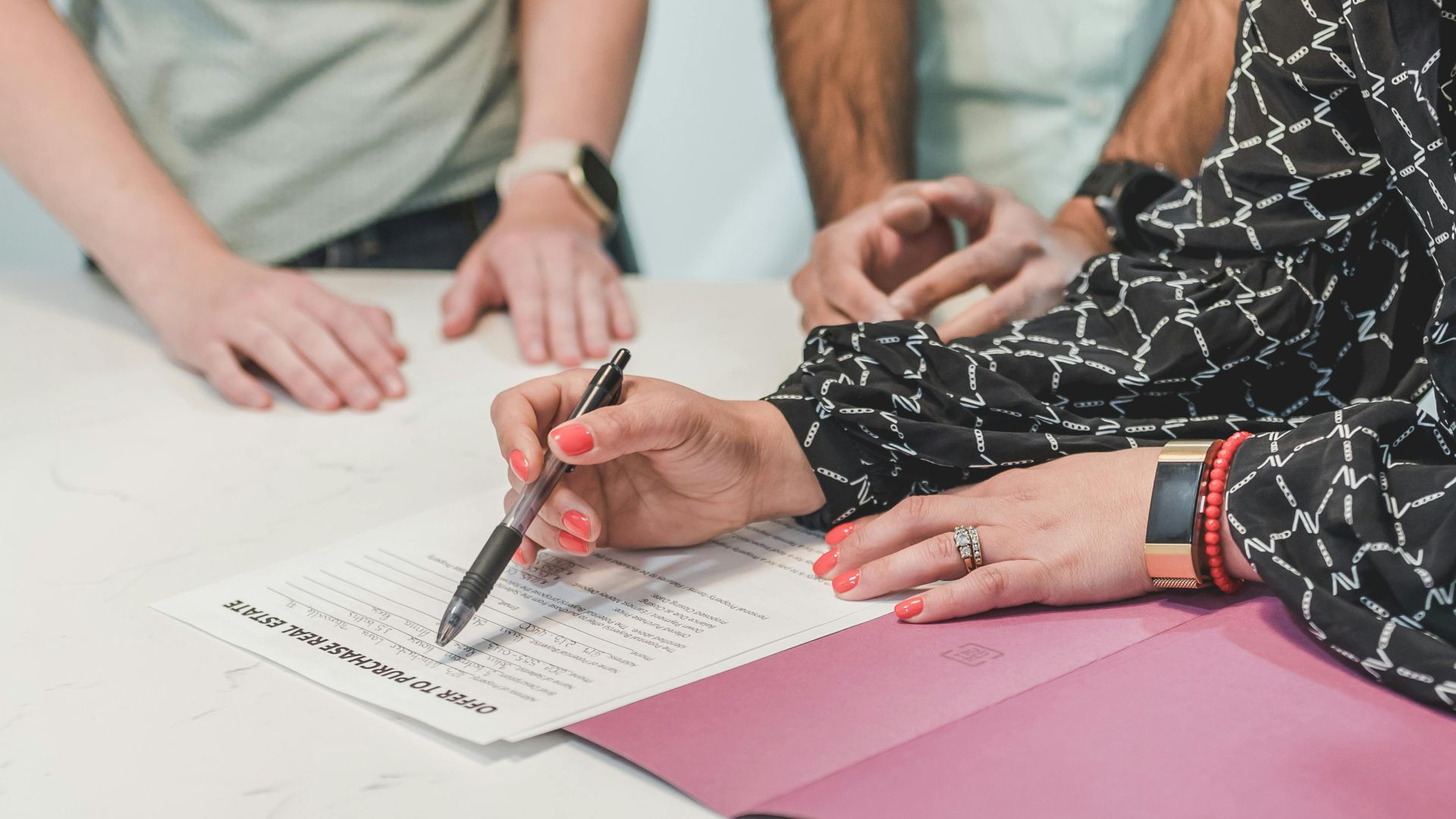 Close-up of hands signing a real estate document at a meeting table with three people.