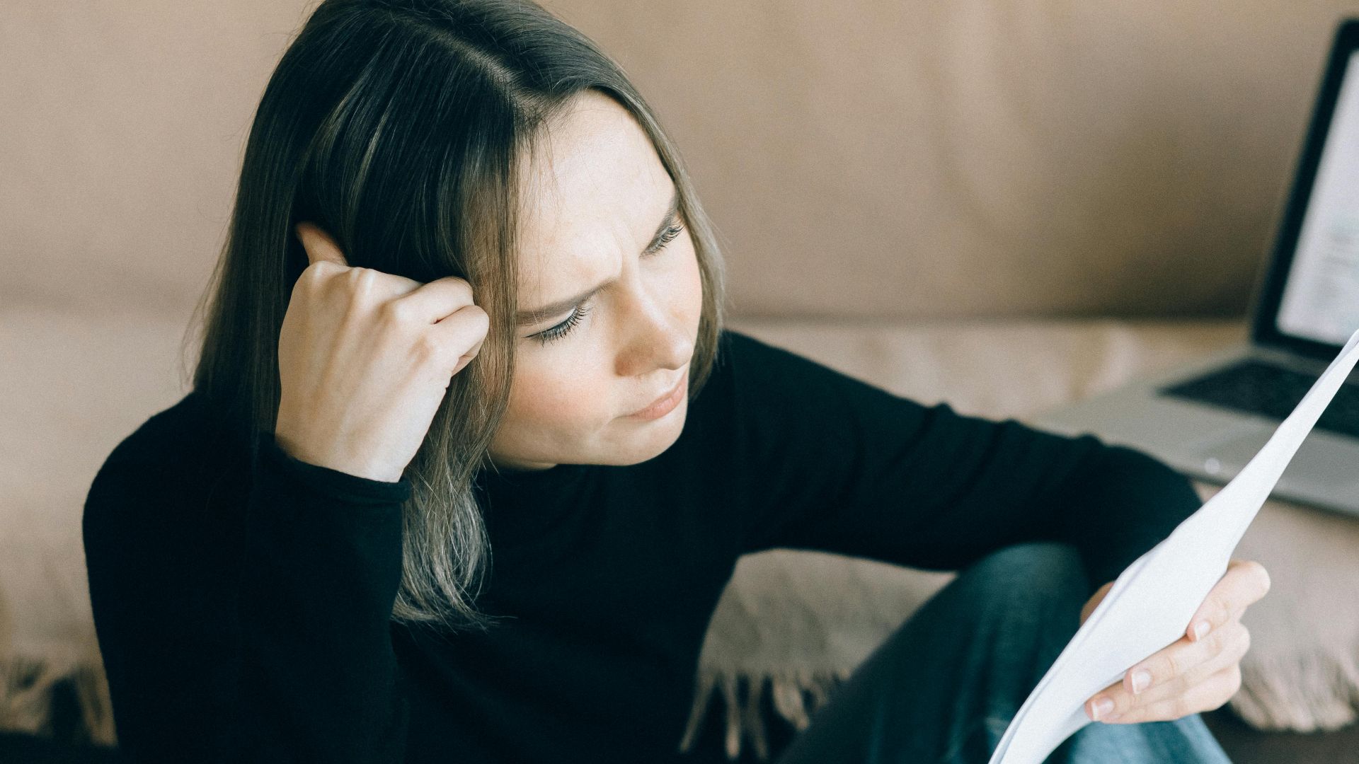 Focused woman analyzing papers with a laptop open, symbolizing thoughtful consideration.