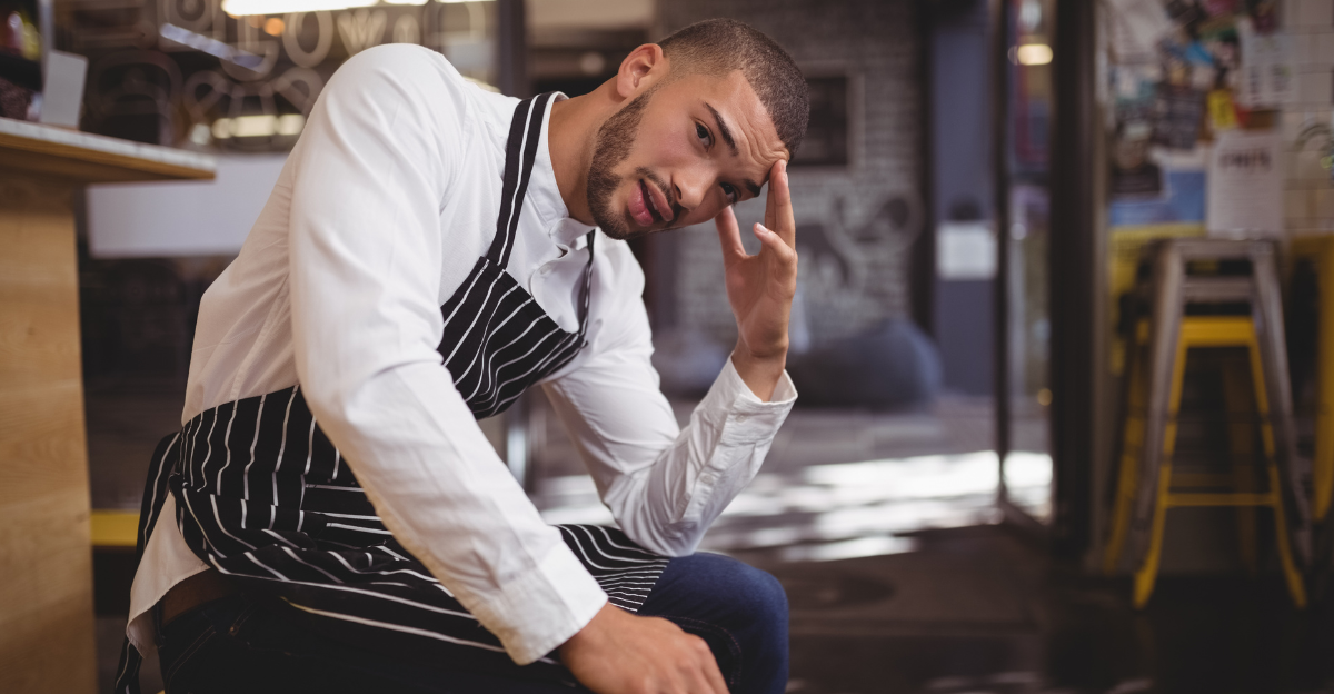 Upset young waiter sitting with headache while looking away at coffee shop