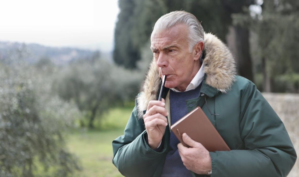 Man in Green Jacket Holding Brown Book and a Pen