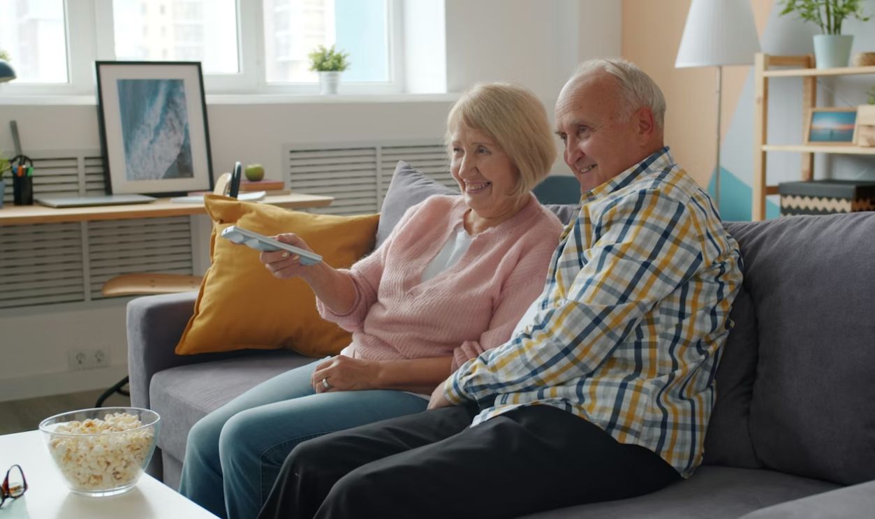 elderly man and woman watching TV laughing talking enjoying movie at home on sofa. Entertainment, lifestyle and leisure concept.