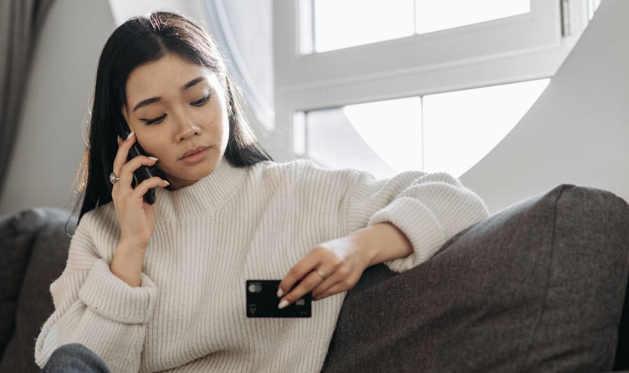 A Woman in Knitted Sweater Talking on the Phone while Holding a Credit Card