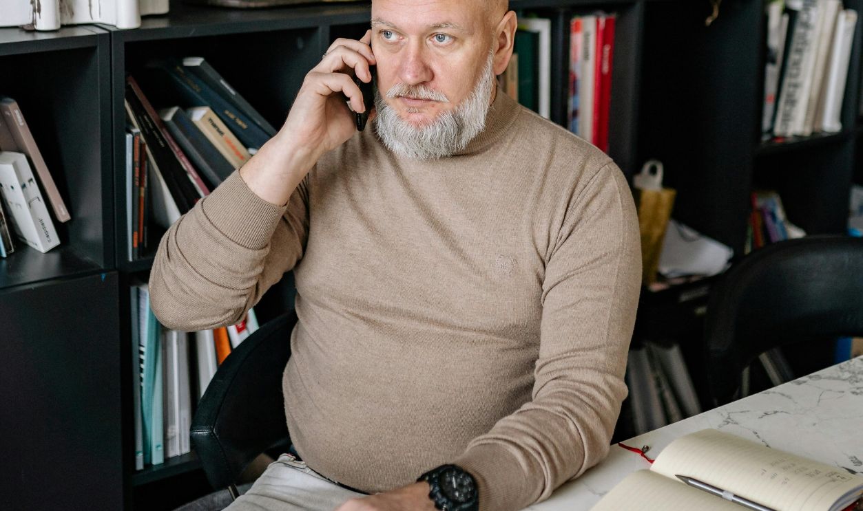 An Elderly Man in Brown Long Sleeves Sitting while Having a Phone Call