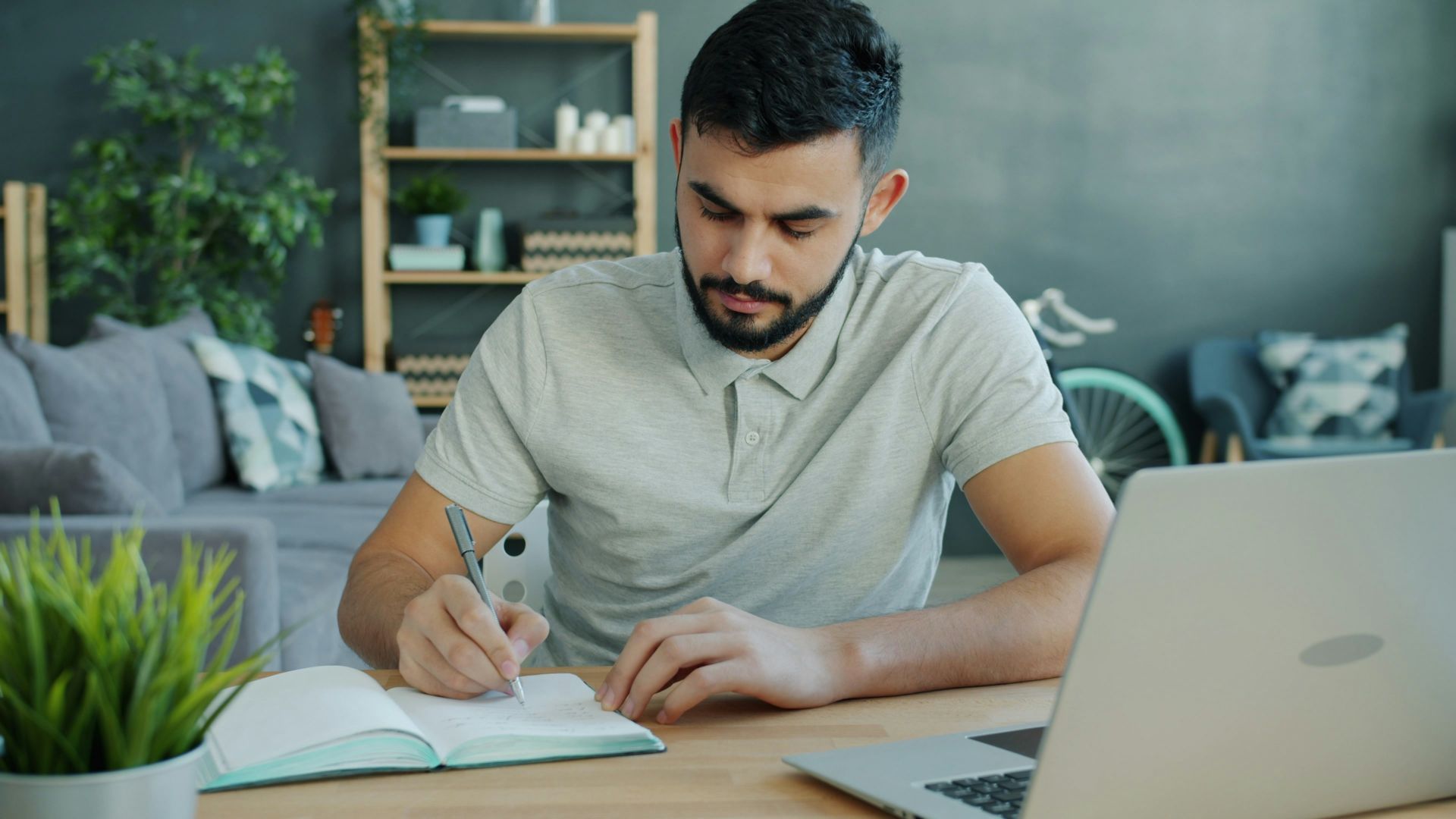 Man writing in notebook at desk with laptop.