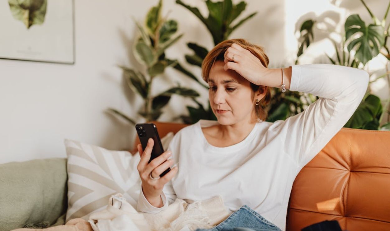 Woman Sitting on Sofa, Looking at Phone and Scratching Her Head