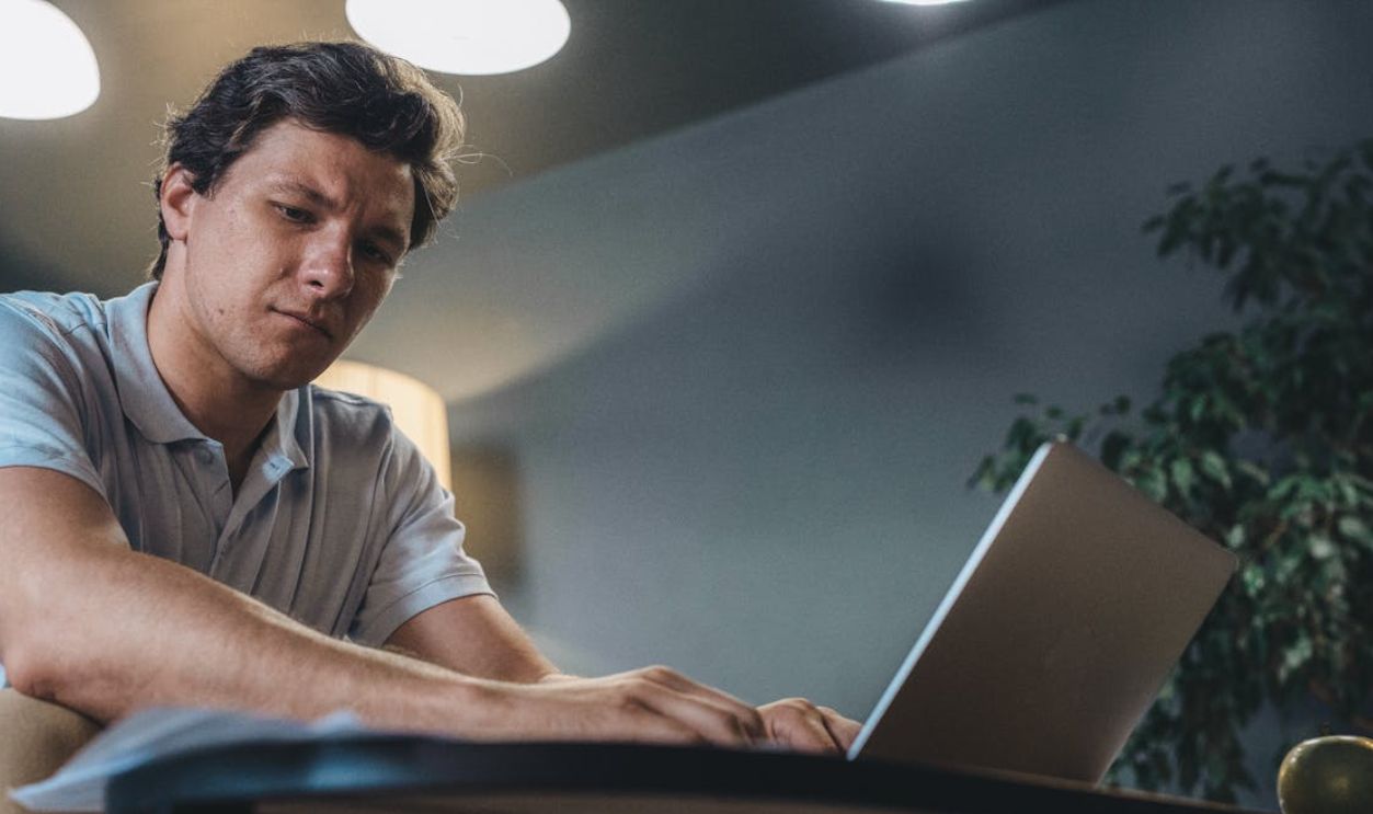 Man in Blue Polo Shirt Looking at the Paper while Typing on the Laptop