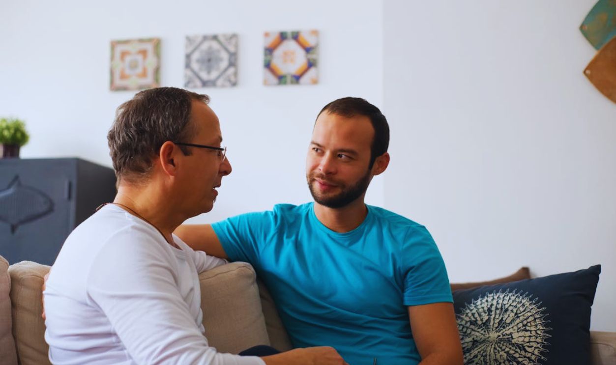 Man in White Long Sleeve Shirt Sitting on Sofa Talking to a Man in Blue Shirt