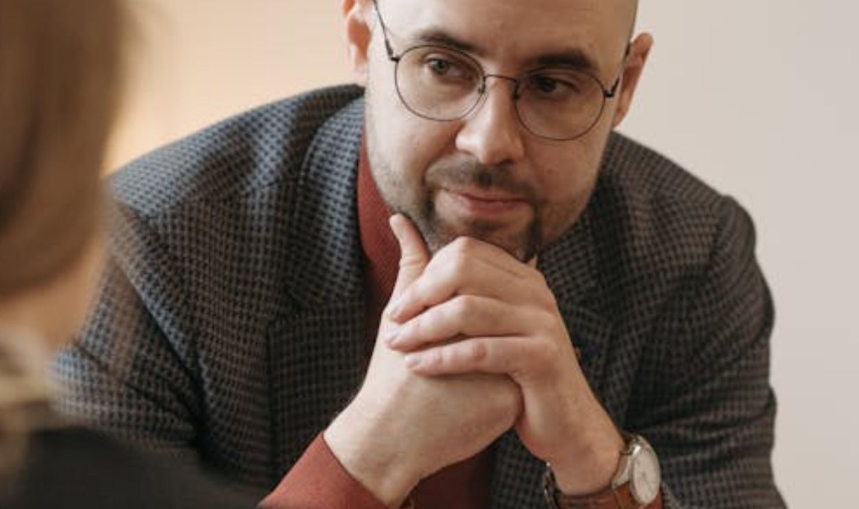 A Man in Gray Suit Jacket and Denim Jeans Sitting with Hands on Chin