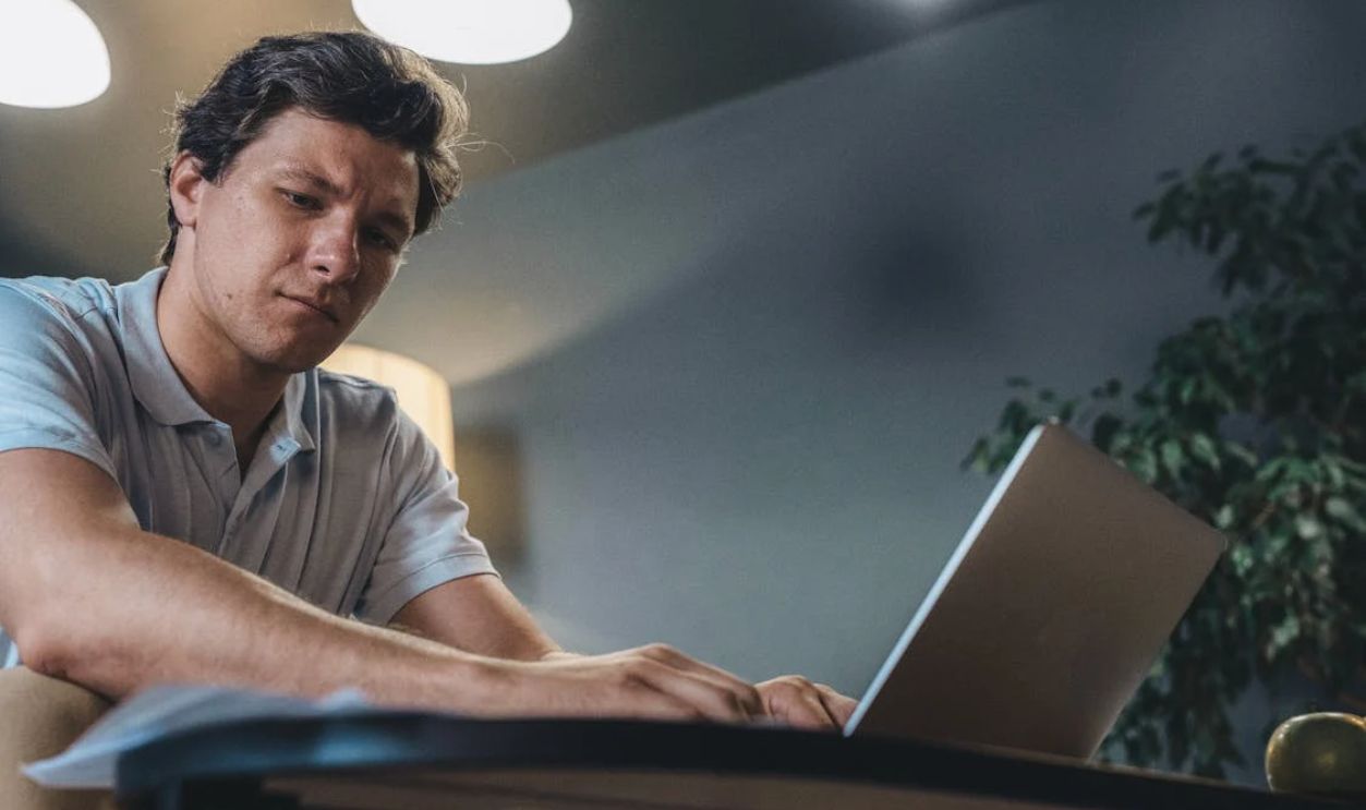 Man in Blue Polo Shirt Looking at the Paper while Typing on the Laptop