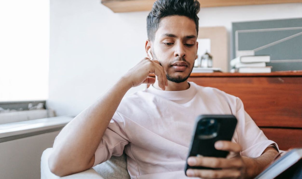 Thoughtful Indian man with wireless earbuds messaging on smartphone