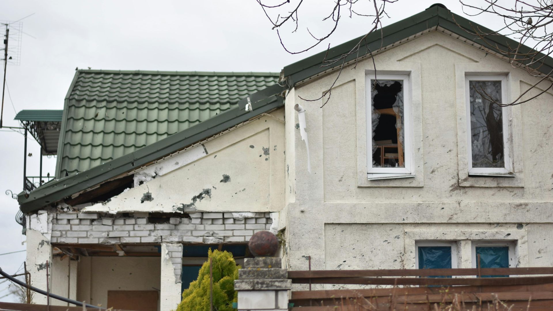 Close-up of an abandoned house with visible damage and a green roof, surrounded by a wooden fence.