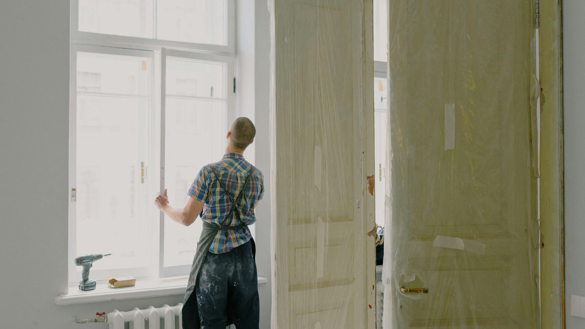 Back view of unrecognizable workman in apron mounting window near drilling machine and doors in flat