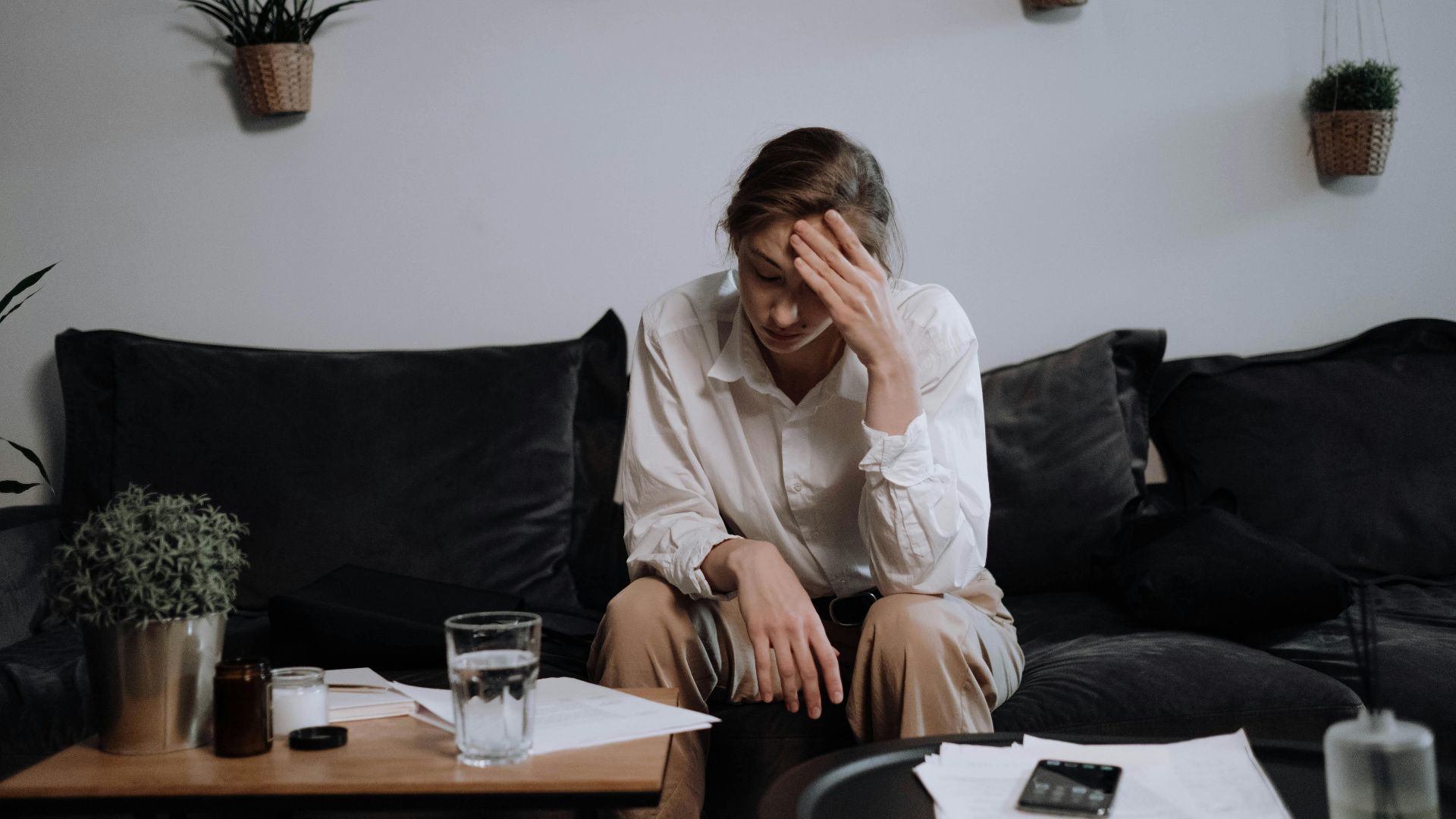 Woman experiencing stress and headache while sitting on a sofa at home, surrounded by papers.