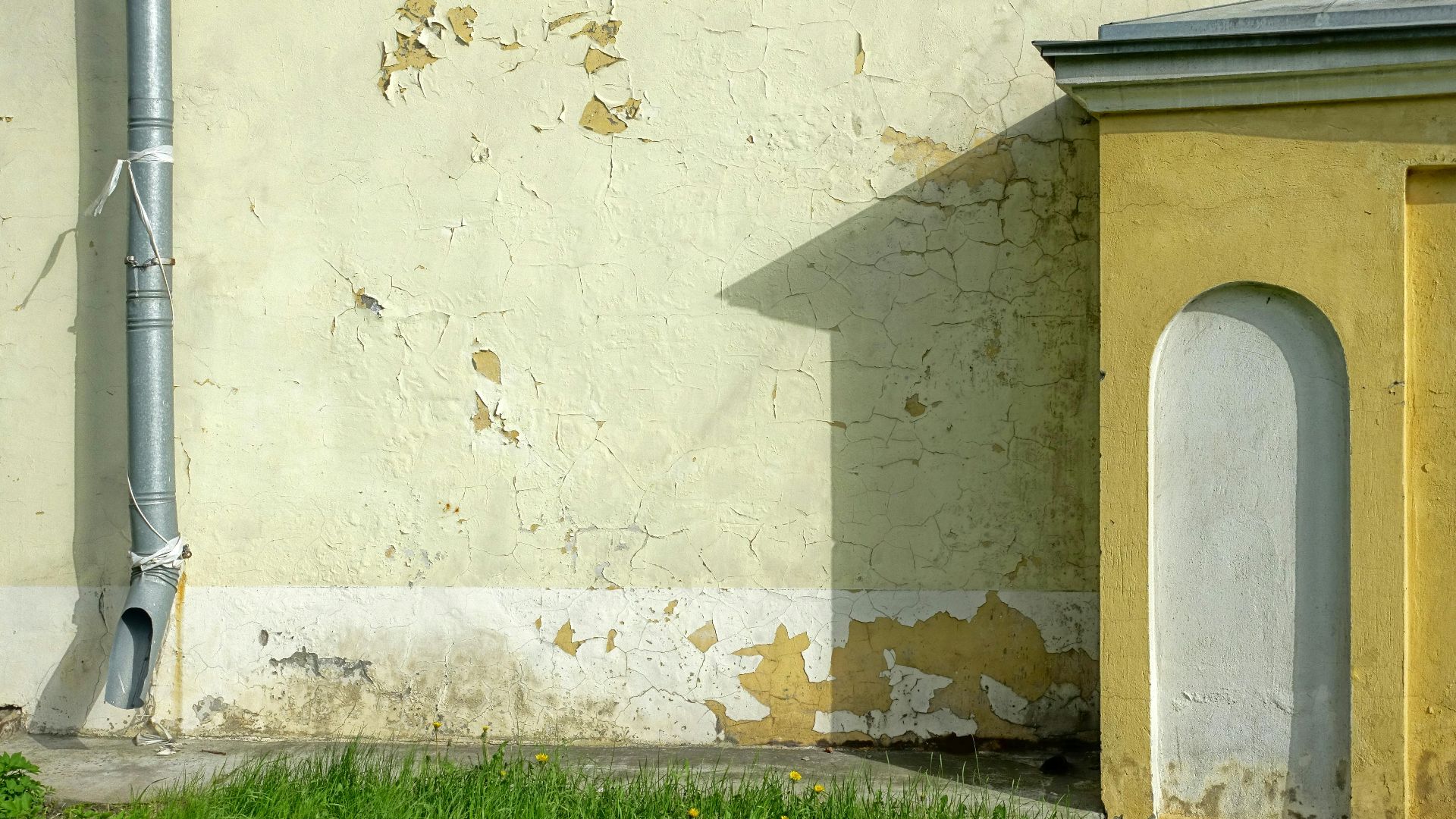Detailed view of an old wall with cracked paint, downpipe, and grass at the base.