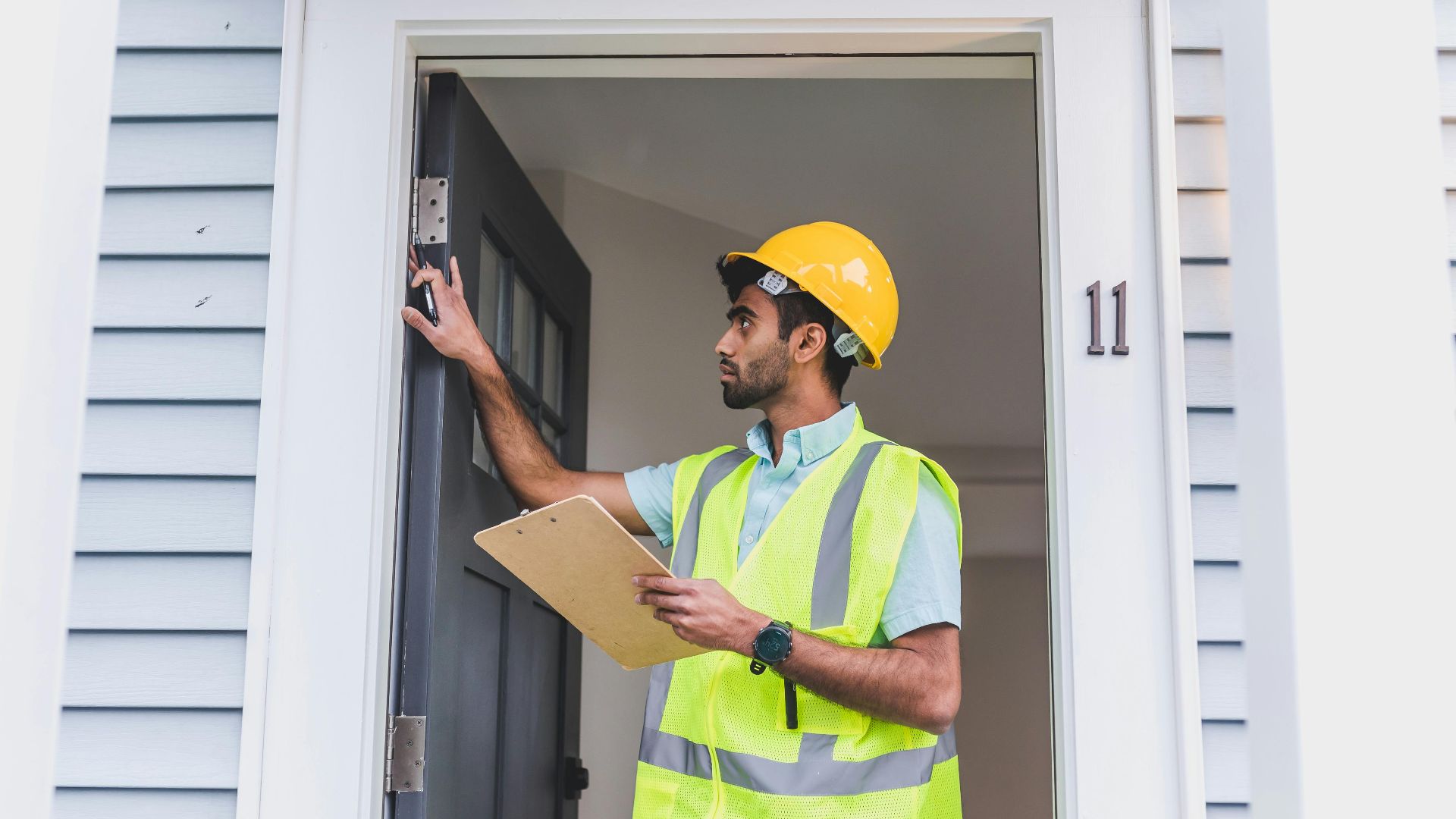 A home inspector in safety vest and hard hat checks doorframe alignment with clipboard outdoors.