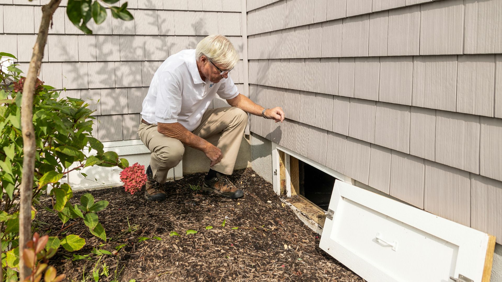 Senior adult inspecting a crawl space entrance during a home inspection on a sunny day.