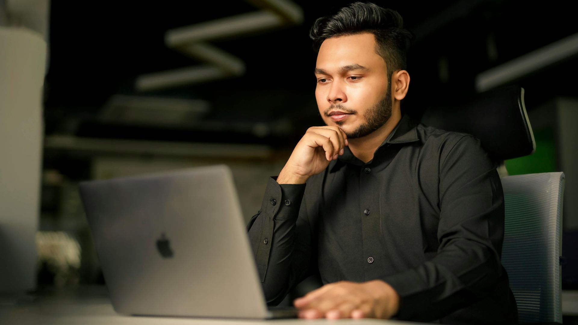 Young South Asian man in black shirt concentrating on work with laptop in modern office.