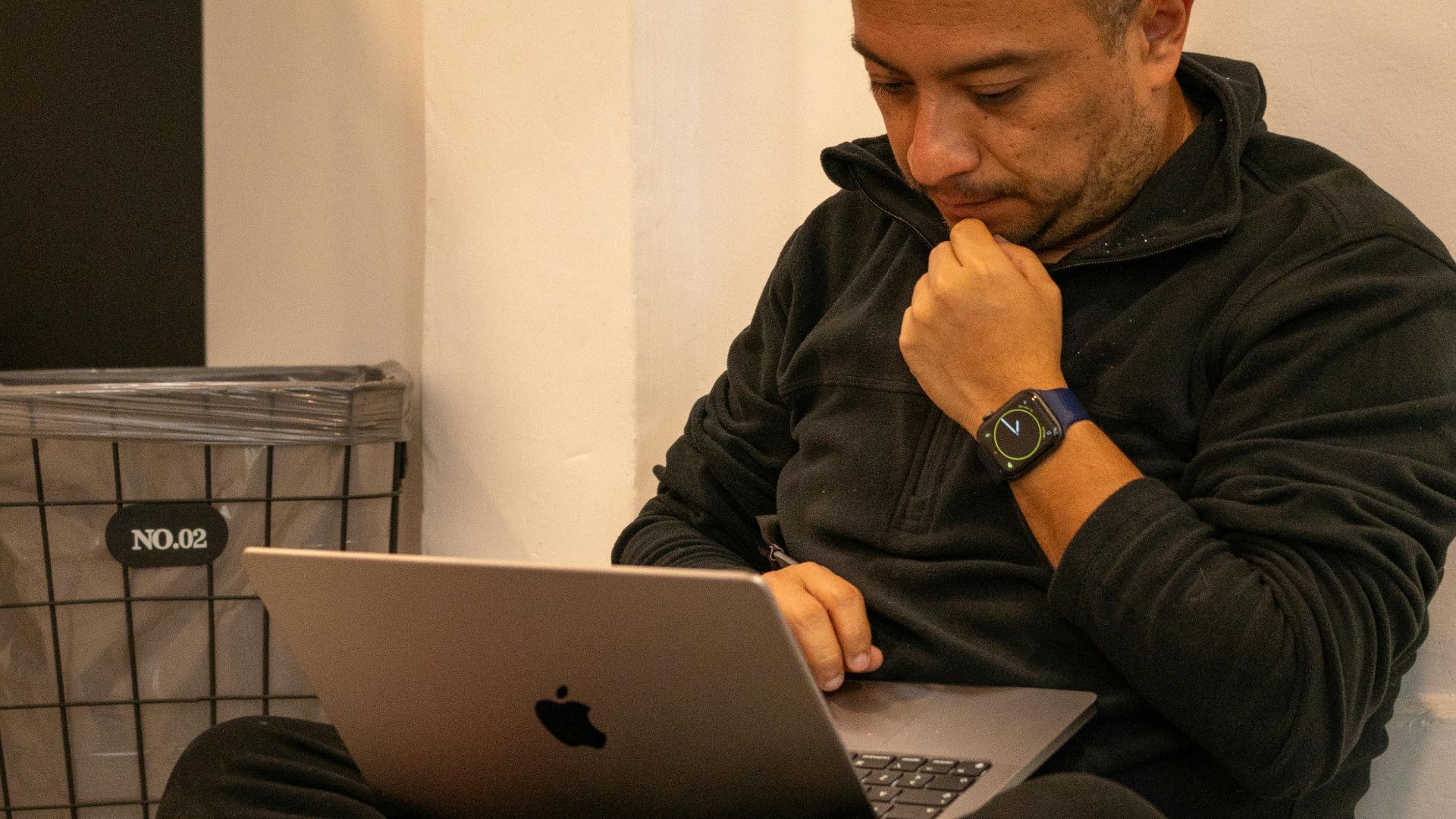 Man sitting cross-legged indoors, focused on his laptop. Casual setting, contemplating or working.