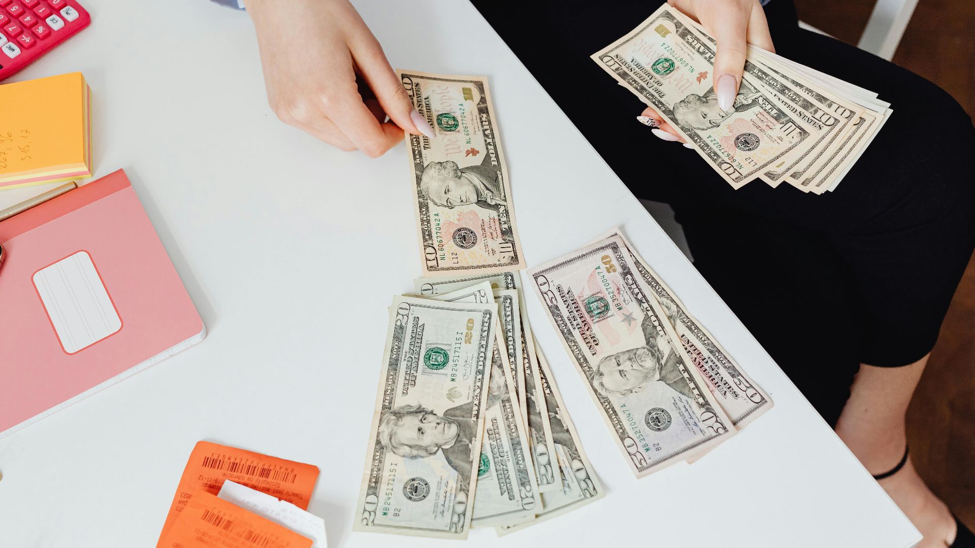 A person counting US dollar bills at a white desk with office supplies nearby.