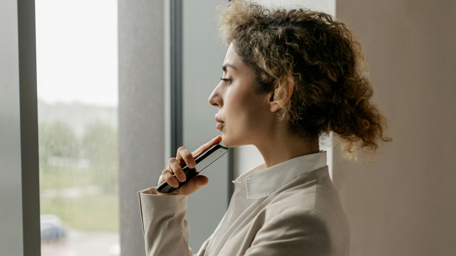 Professional woman in suit looking thoughtfully out a glass window, suggesting corporate environment.