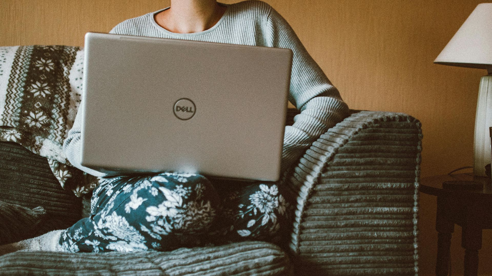A woman in casual clothing works on a laptop while relaxing on a cozy sofa indoors.