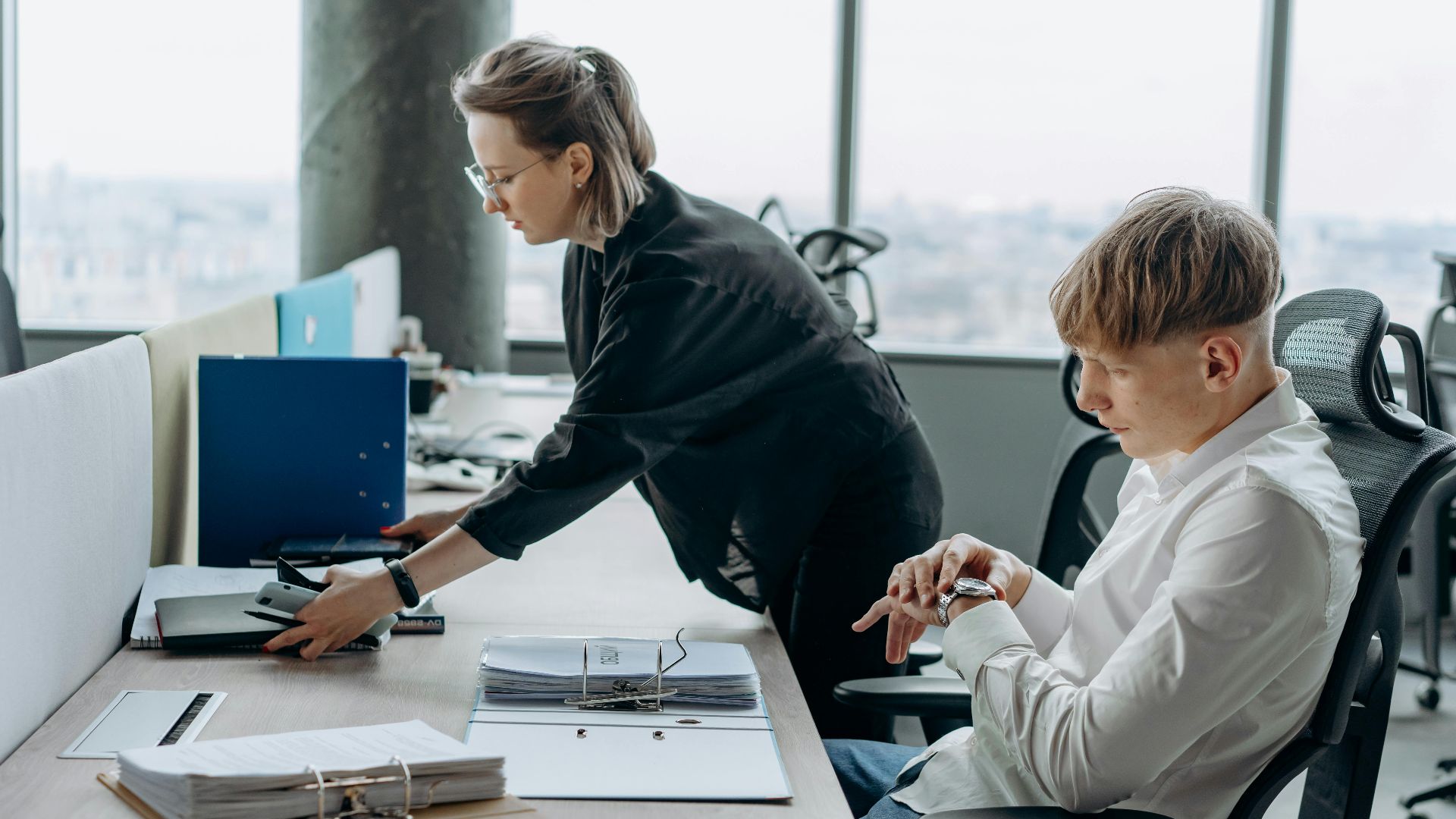 Two business professionals working together in a modern office environment with documents and computers.