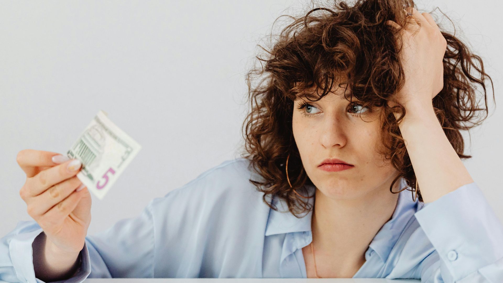 Concerned woman with curly hair holding a five dollar bill, expressing financial worry.