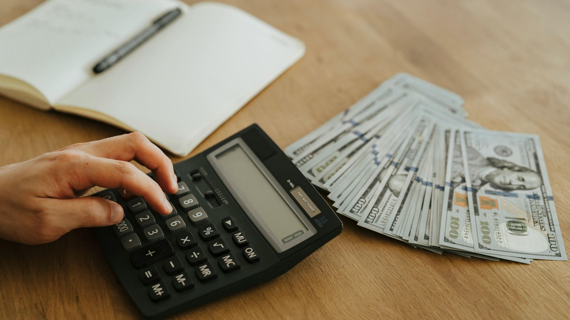 Close-up of a hand using a calculator with cash and a notebook on a wooden table.