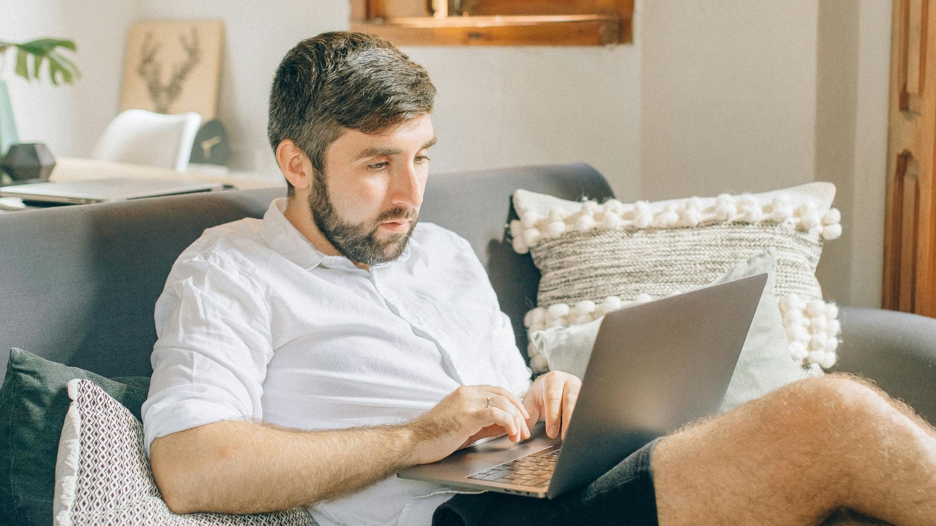 Adult man sitting on couch using laptop, working remotely from cozy home setting.