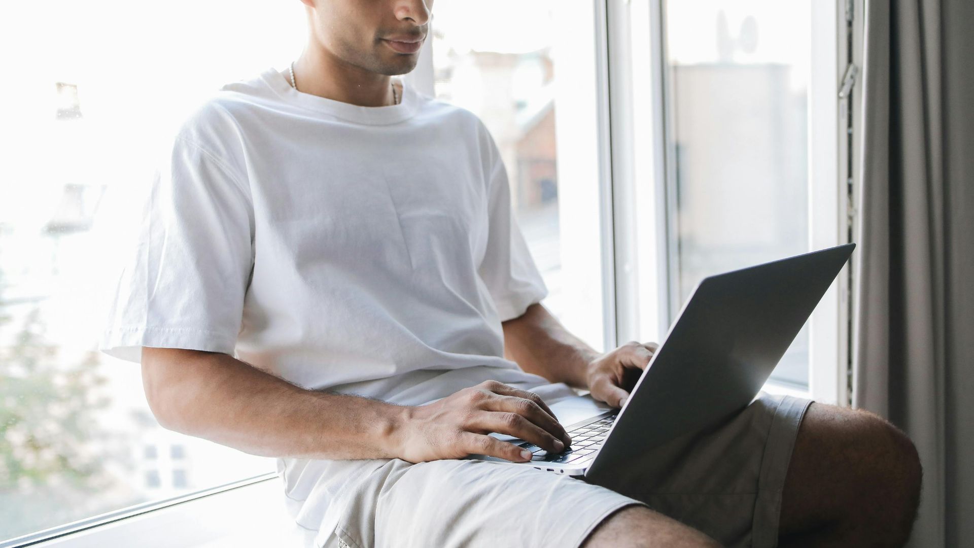 A man in a white t-shirt uses a laptop while sitting by a window, focusing on work or study.