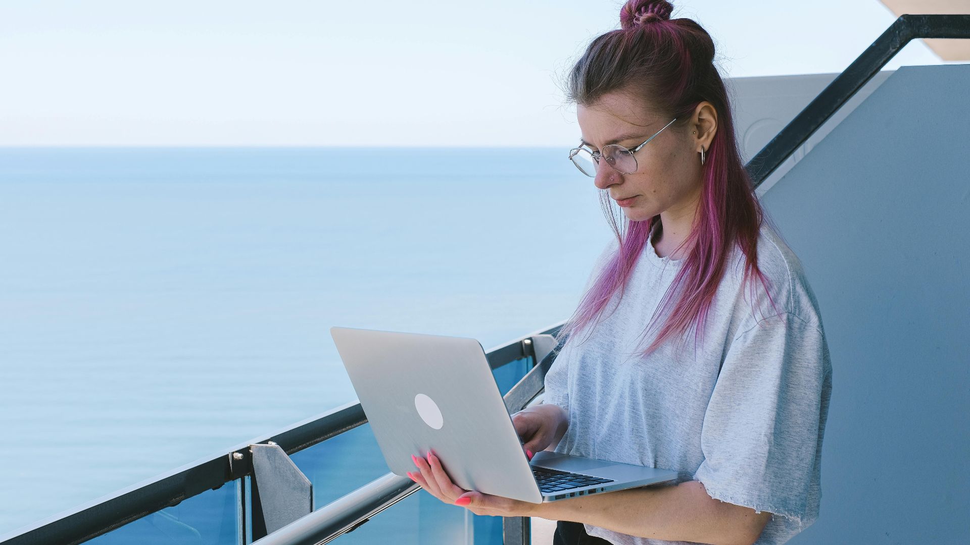 A young woman works remotely on her laptop by the sea on a sunny day, embodying the digital nomad lifestyle.