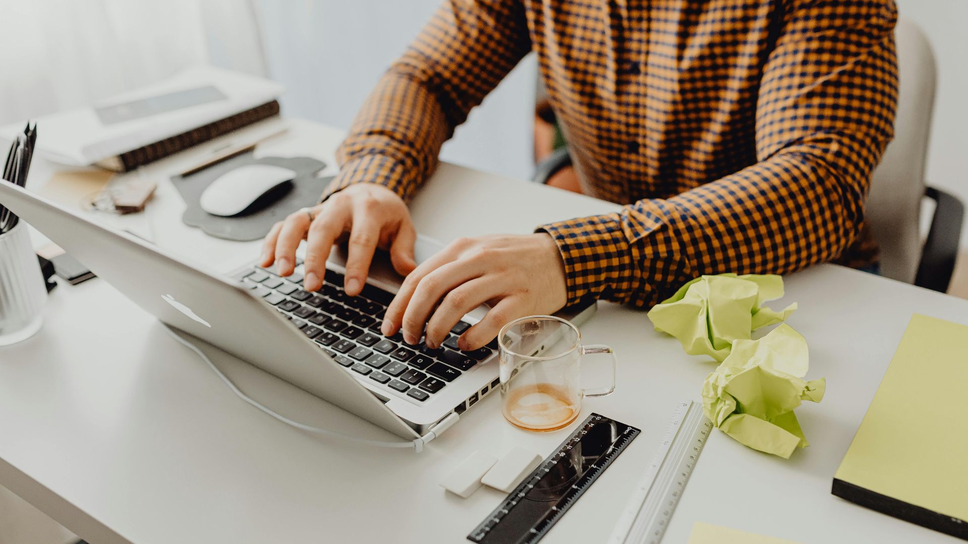 A man wearing a checkered shirt types on a laptop at his office desk.