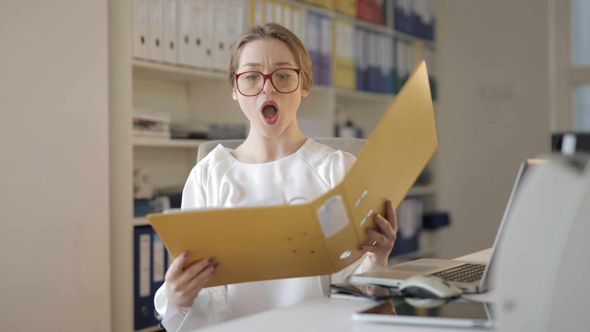 Surprised woman with eyeglasses reading a file in an office filled with shelves of records.