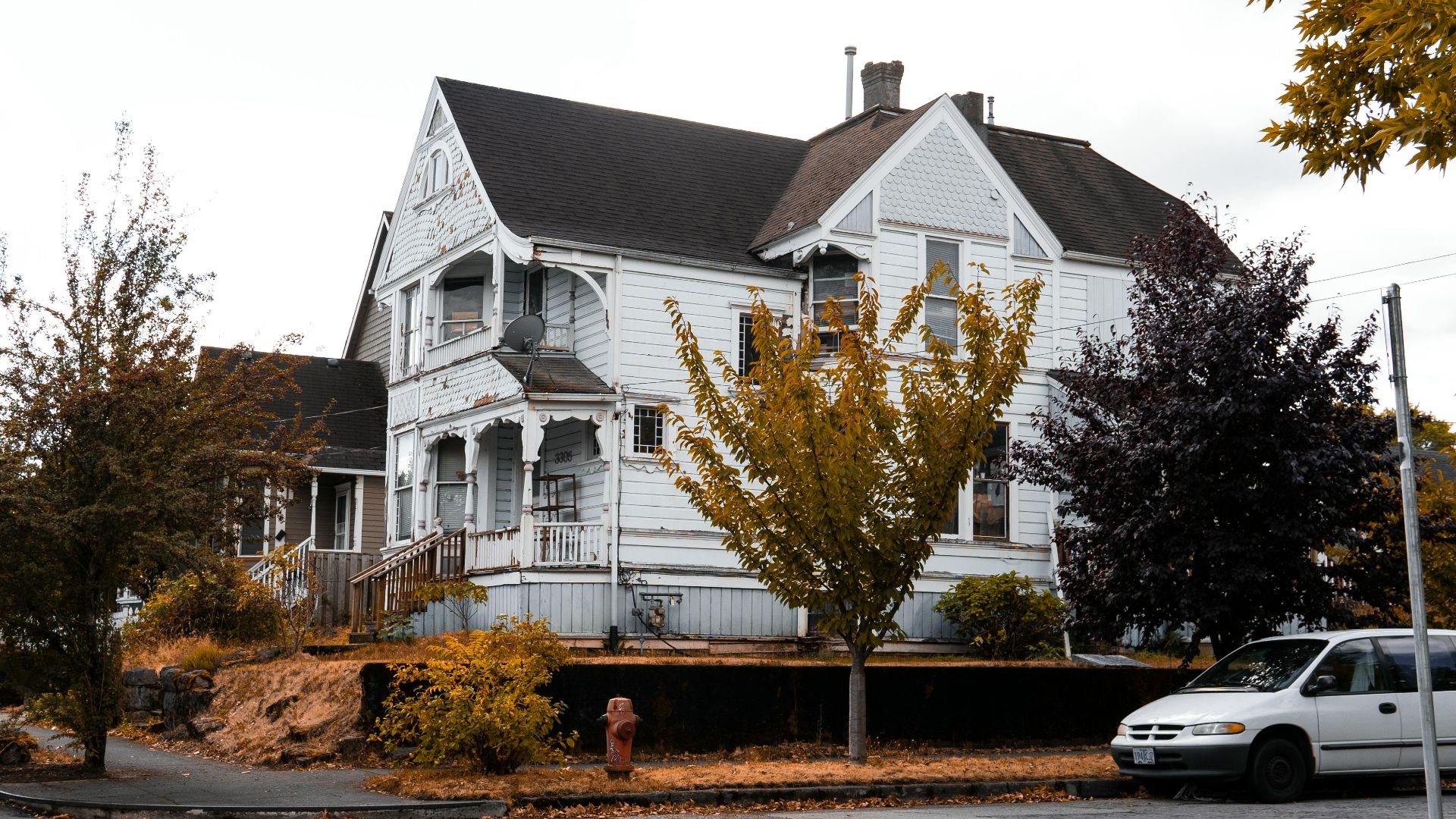 A weathered white wooden house with autumn trees, parked car, and fall ambiance on a suburban street.
