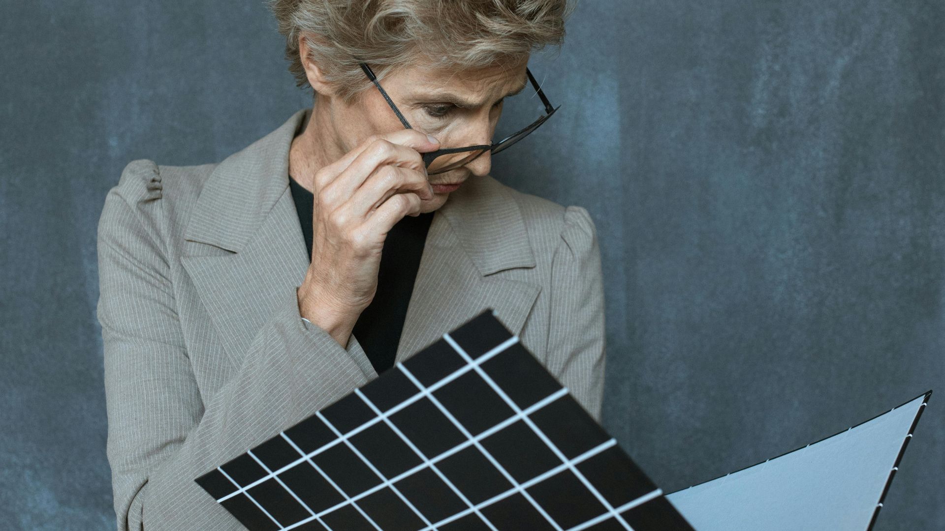 Elderly woman with glasses studies a black portfolio against a gray wall.