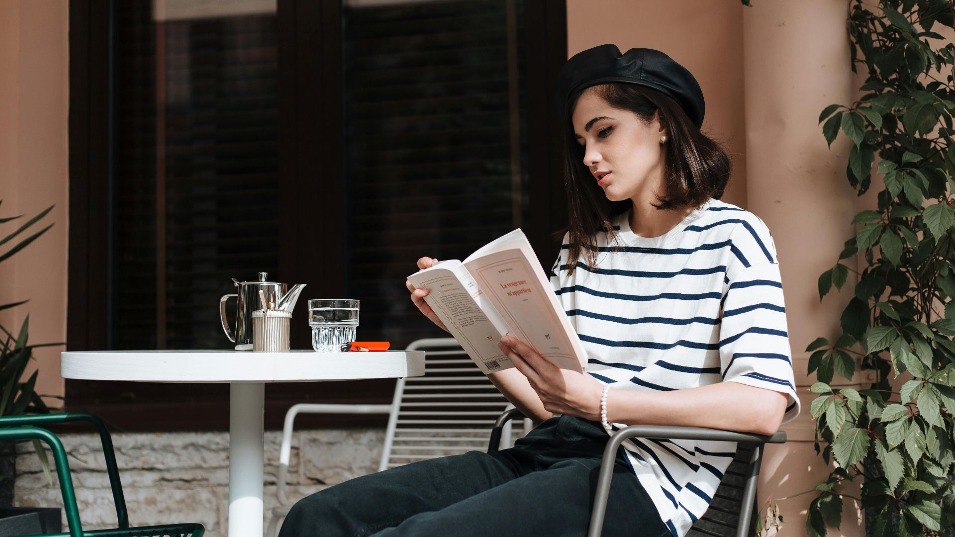 Casual outdoor scene of a woman reading at a cafe, wearing a striped shirt and beret.