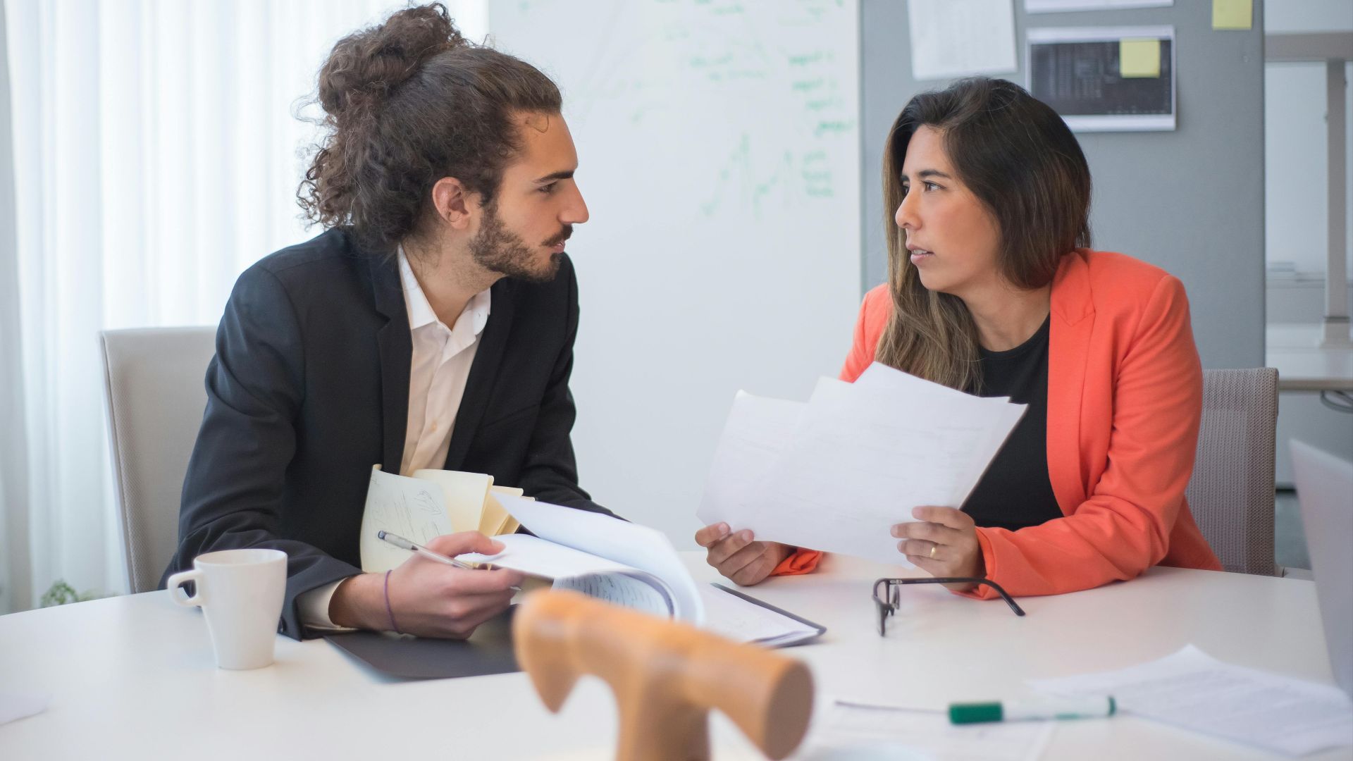 Two colleagues engaged in a discussion at an office table with documents and coffee.