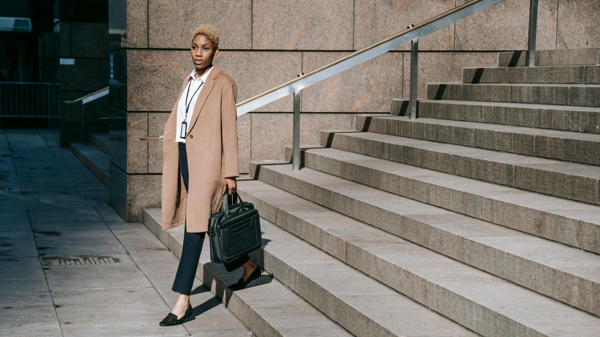 Stylish young African American businesswoman with short blond hair in elegant outfit walking downstairs of modern building after workday
